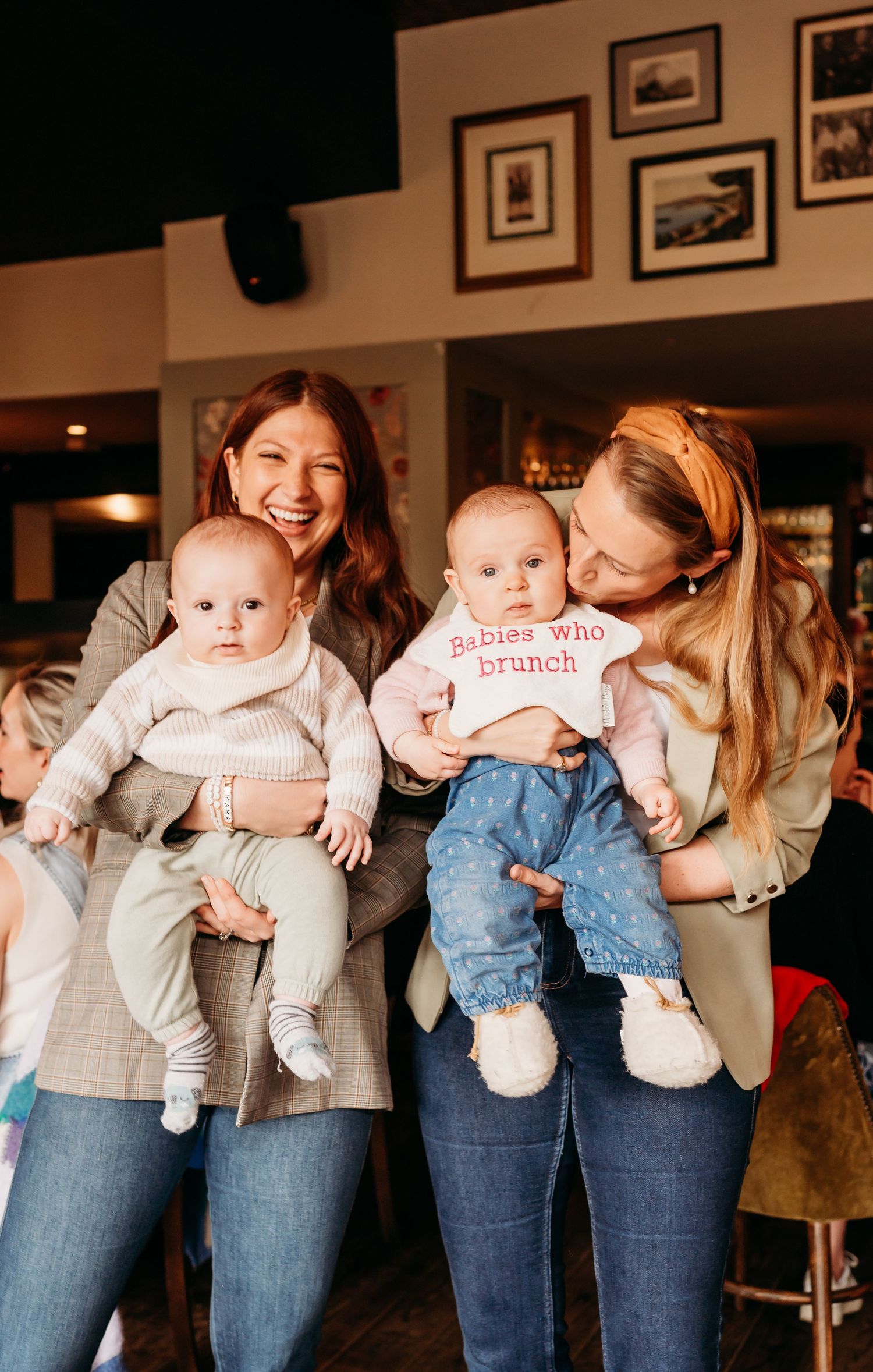 Four babies in matching white outfits held by women wearing jeans at a brunch gathering.