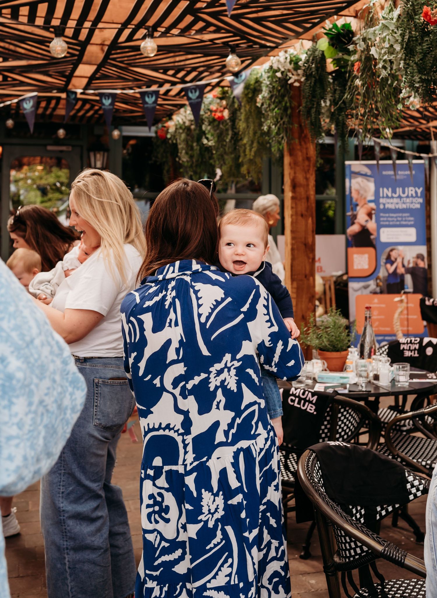 People mingle at an outdoor market or event space decorated with string lights and greenery under a striped canopy.