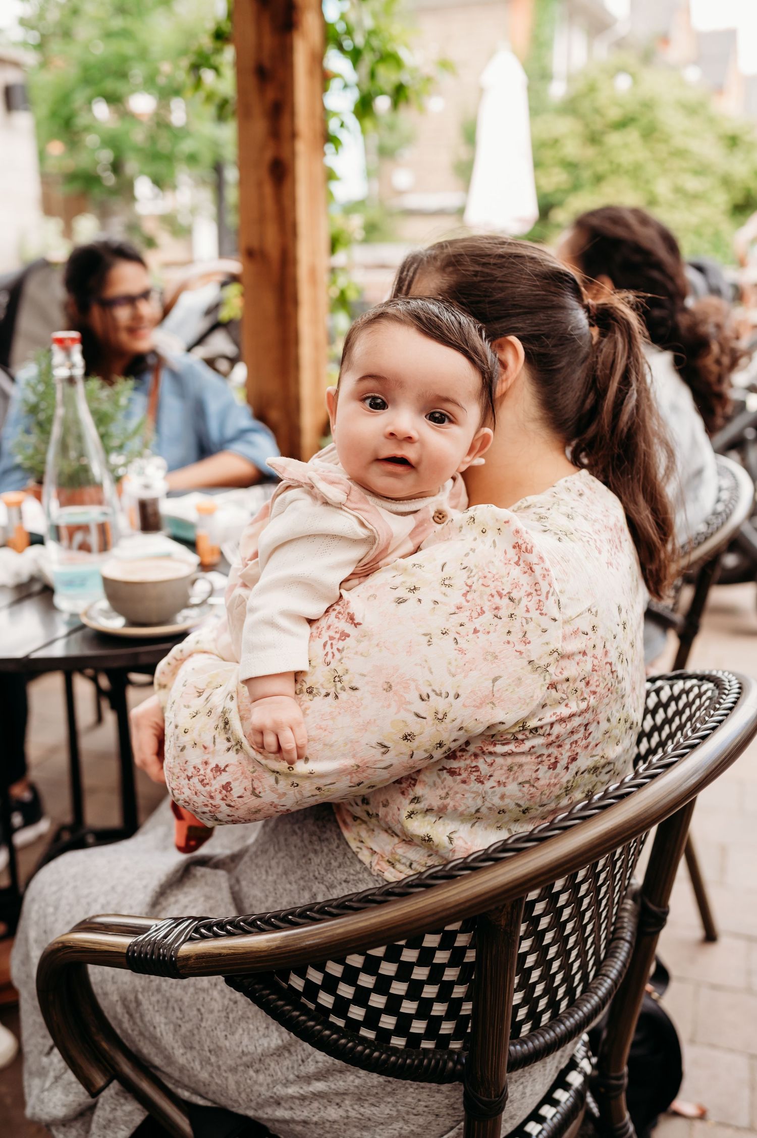 A baby in a floral pink dress lies back in a metal patio chair while someone leans close during an outdoor gathering.