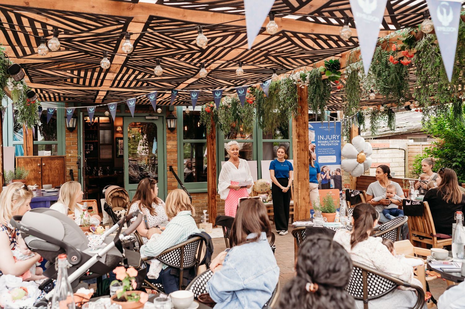 A crowded outdoor restaurant patio with wooden pergola, white banners, and lush greenery on a sunny afternoon.