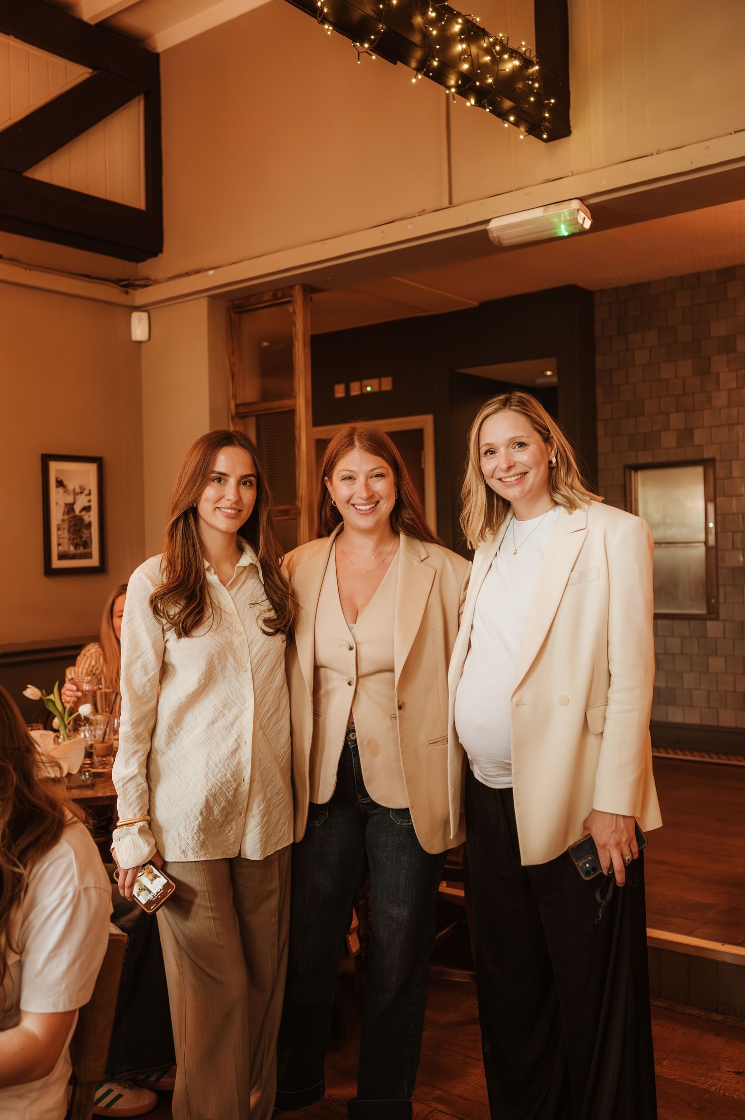 Three women pose together wearing cream and neutral toned business attire in a warmly lit room with string lights.