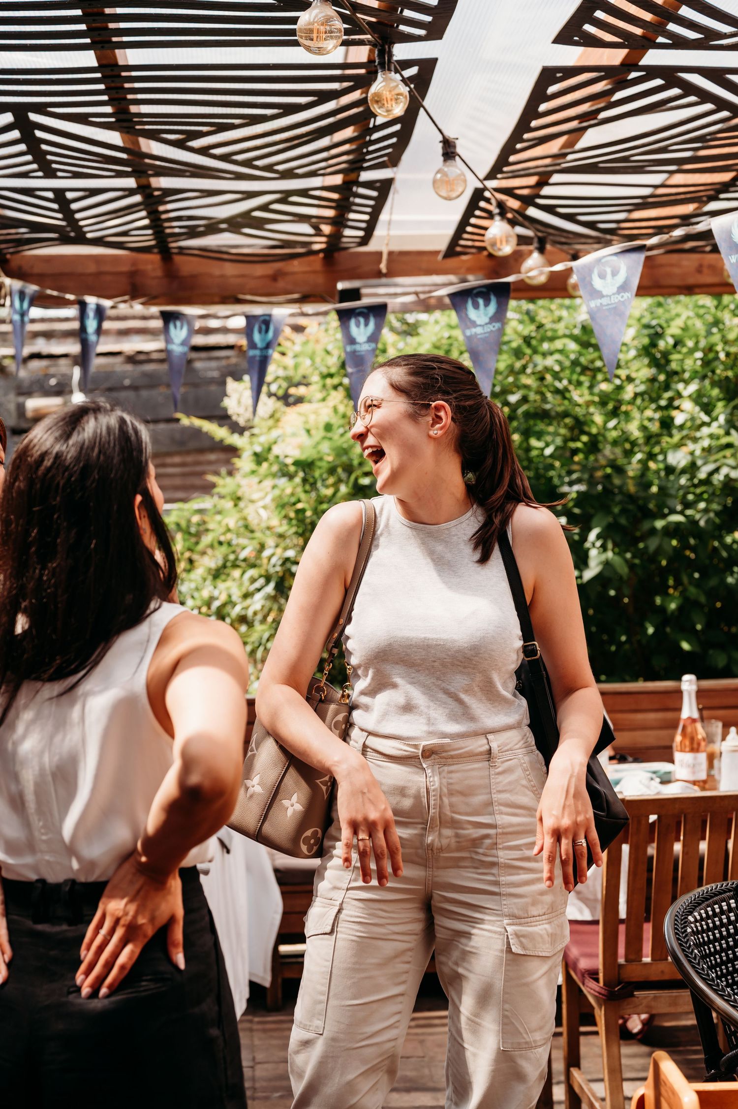 Two people chatting and laughing at an outdoor patio with wooden beams and string lights overhead.