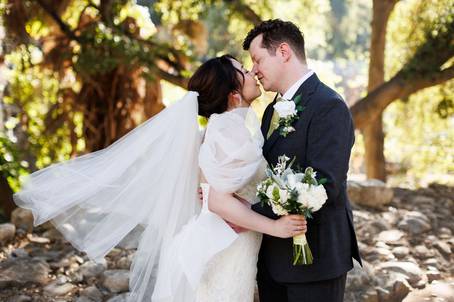 A romantic wedding couple embraces under dappled sunlight in a garden setting, with the bride's veil flowing in the breeze.