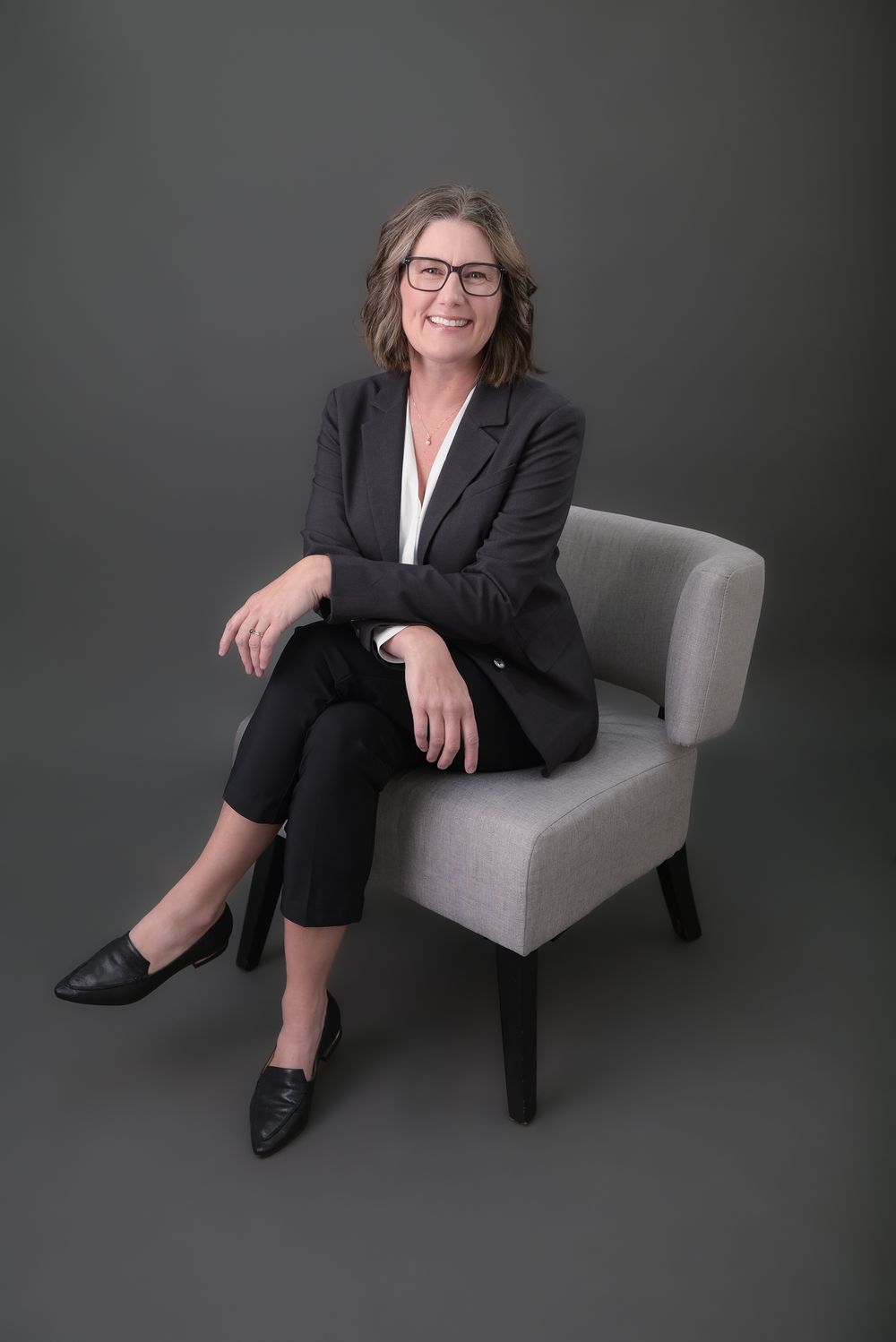 Professional business portraits in a black suit seated in a grey chair.