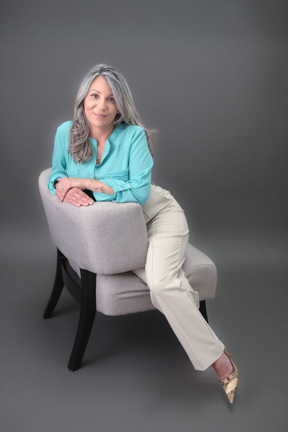 Studio portraits in a turquoise top lounging in a grey modern chair.
