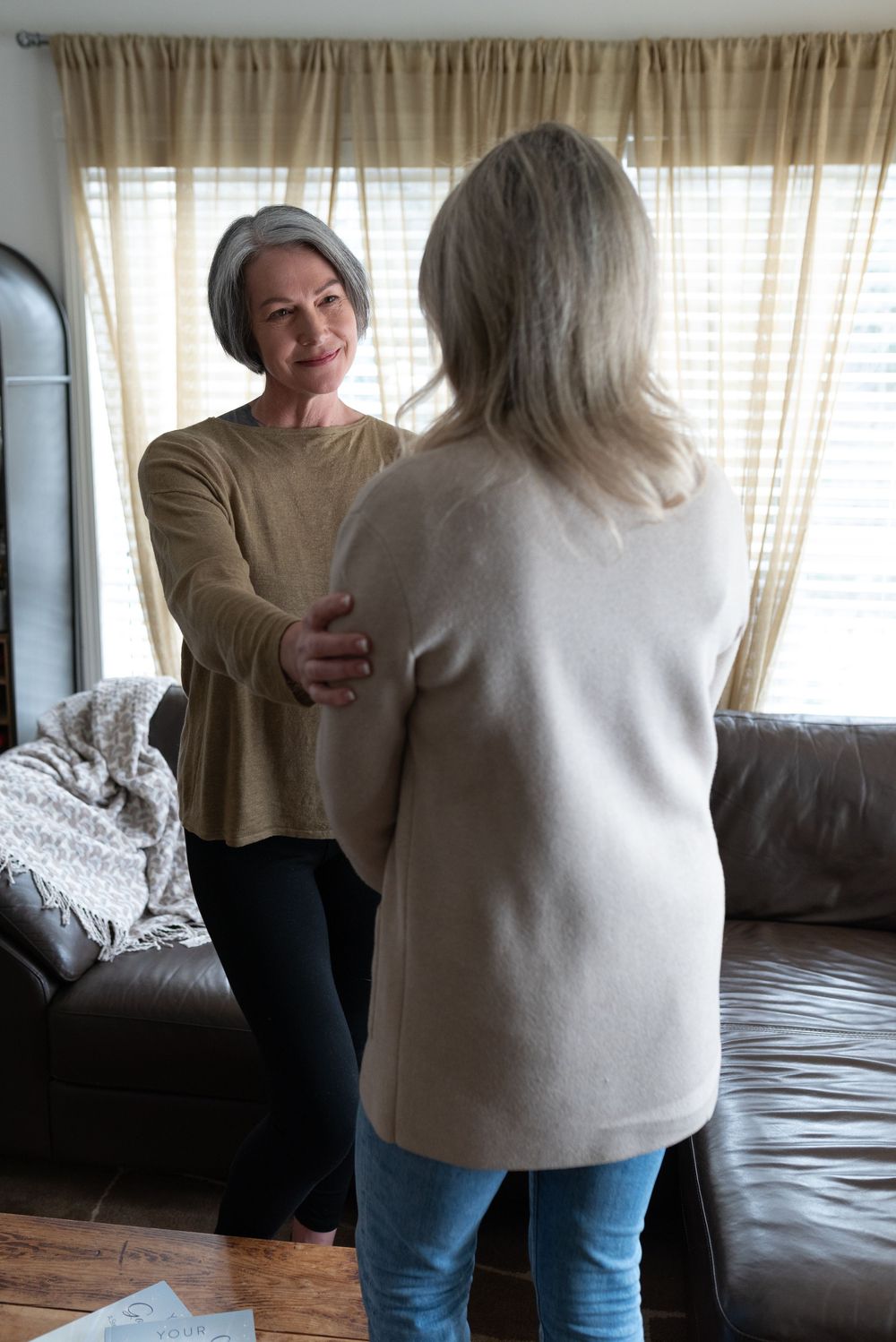 Two people share an emotional embrace in a living room setting with curtained windows and a couch visible.