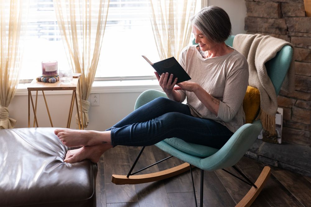 A person in casual attire sits comfortably in a modern teal accent chair while reading near a window.