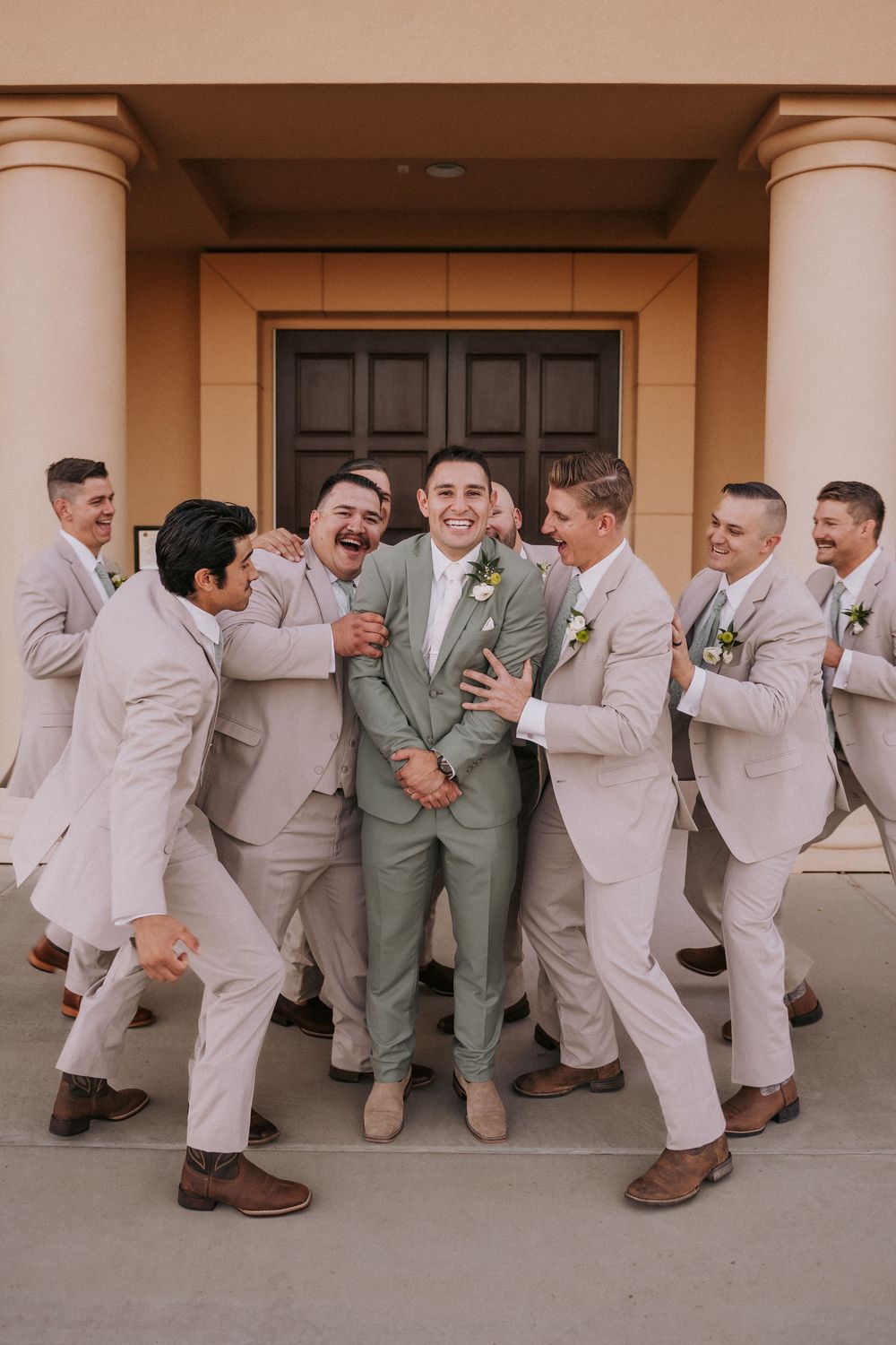 Groomsmen playfully pose with the groom in front of a columned entrance. Affordable wedding photographers in New Mexico. Queer elopement NM