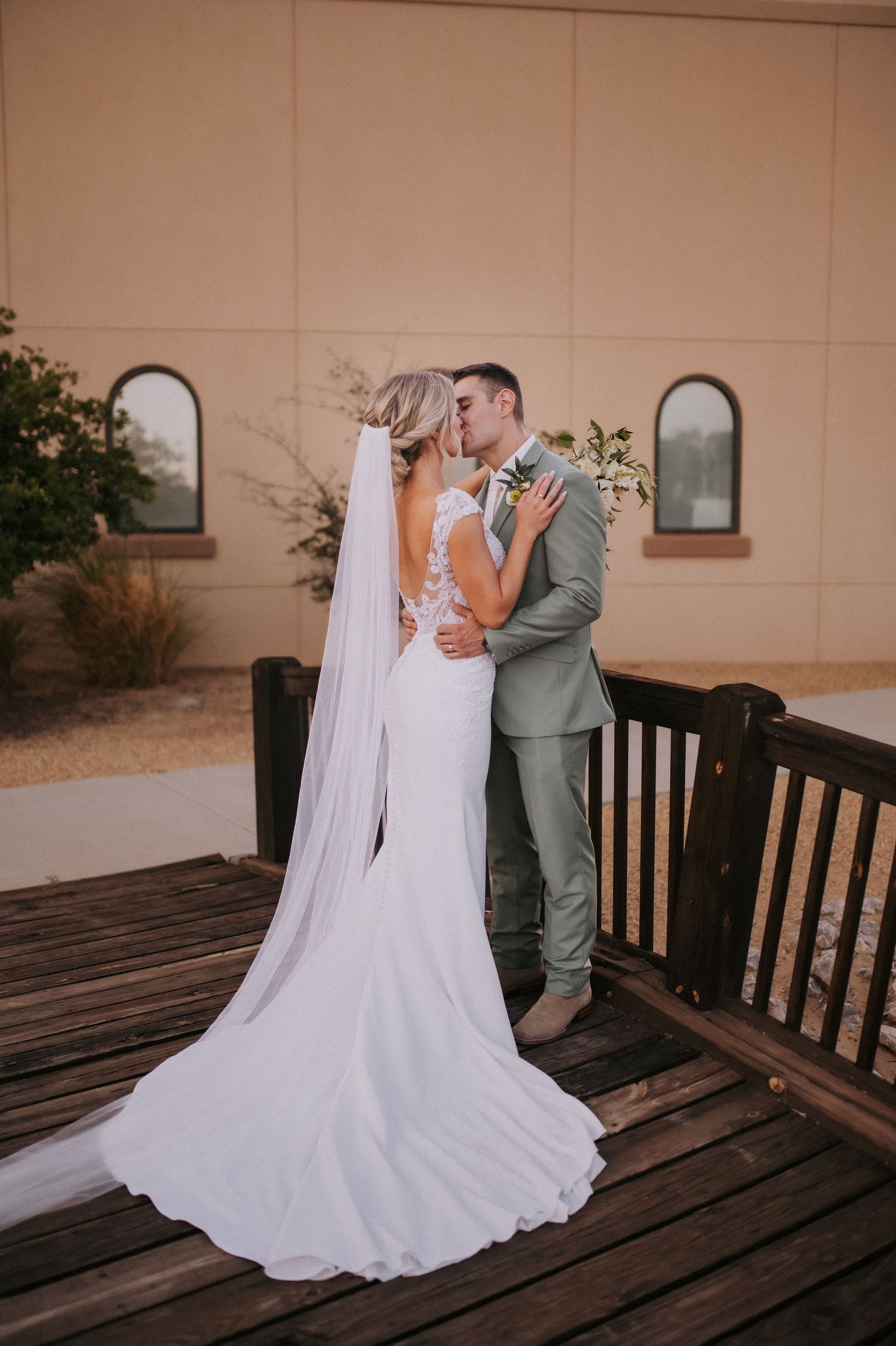 A wedding couple embraces against a beige building with arched windows and a flowing white bridal veil. Queer wedding photographer in ABQ
