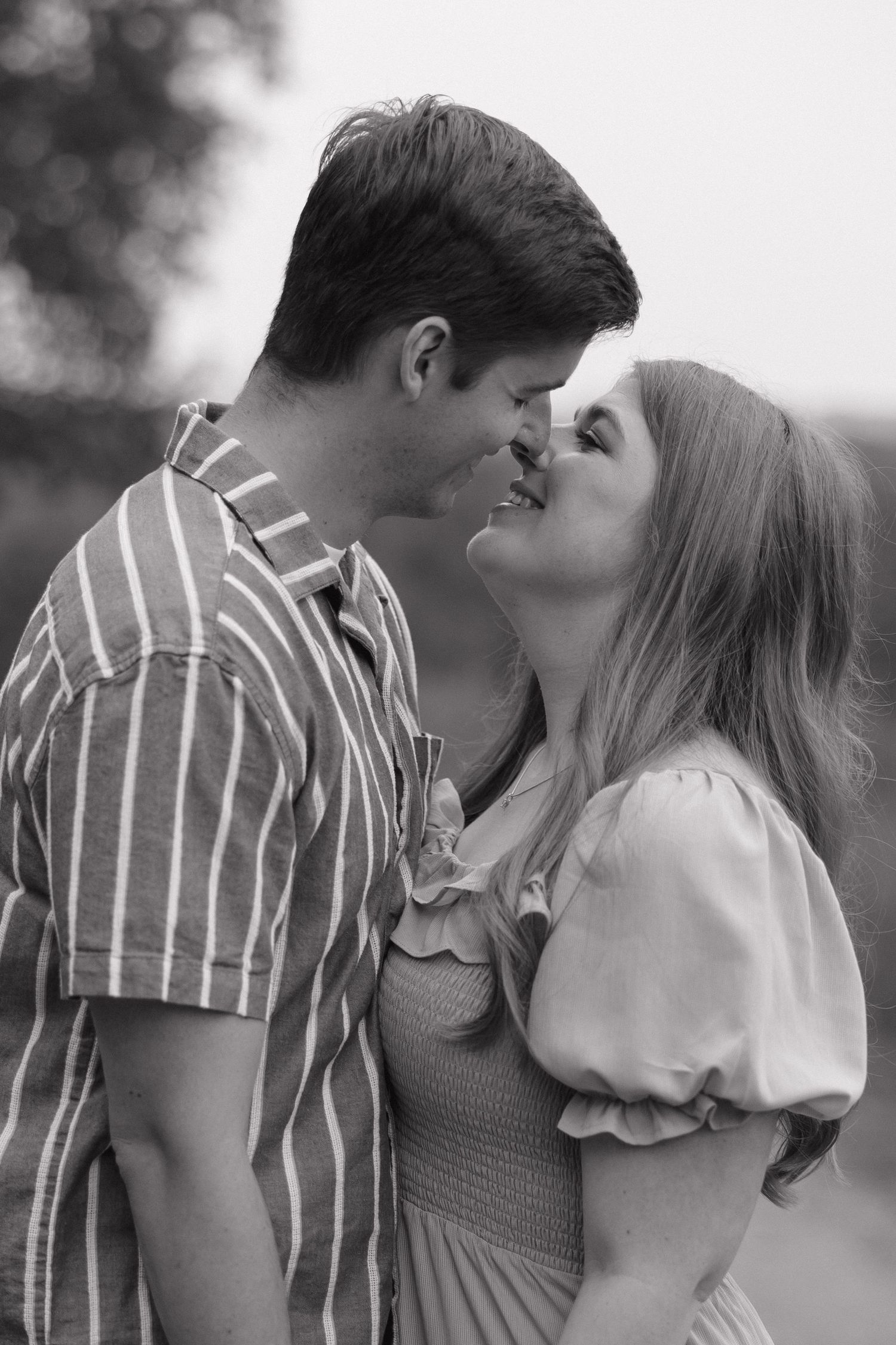 A romantic couple share an intimate kiss outdoors in black and white, wearing a striped shirt and ruffled blouse.
