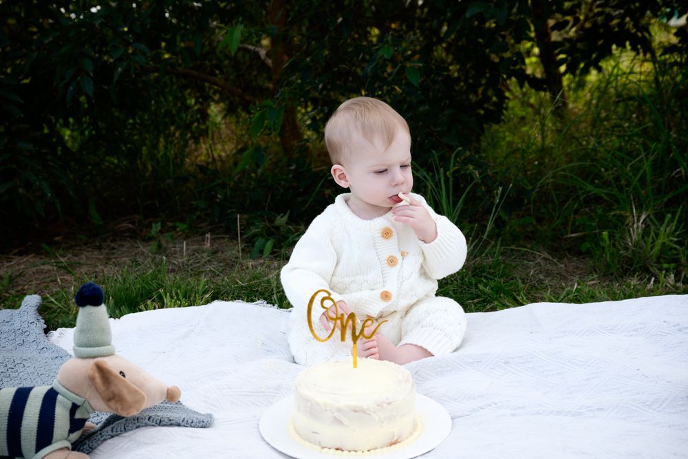 A baby wearing a white outfit sits with a decorative cake in an outdoor setting at a one year birthday celebration.