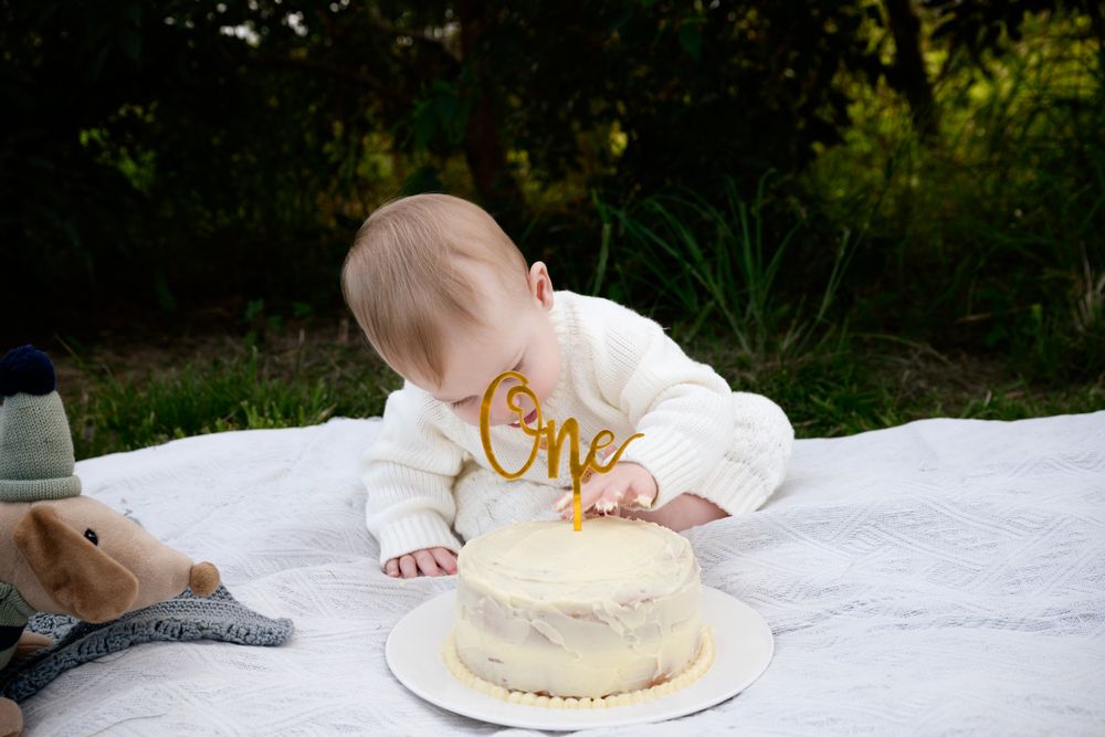 A baby in white sits on a blanket with birthday cake and smash cake decorations at a first birthday photoshoot.