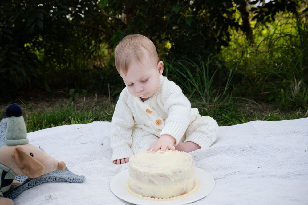 Baby wearing white outfit reaching towards birthday cake on white blanket outdoors during cake smash photoshoot.