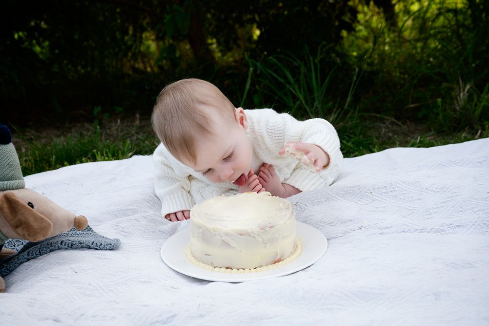 Baby seated on white blanket outdoors excitedly reaching for cake during first birthday celebration.