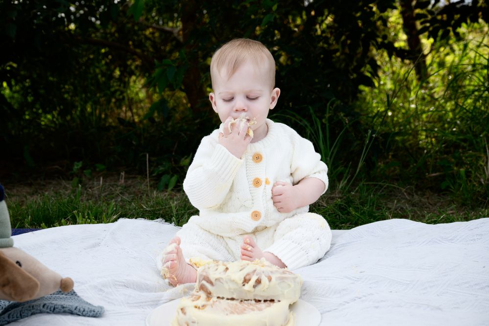 Baby in white outfit sitting on blanket enjoying cake during outdoor smash cake photo session.