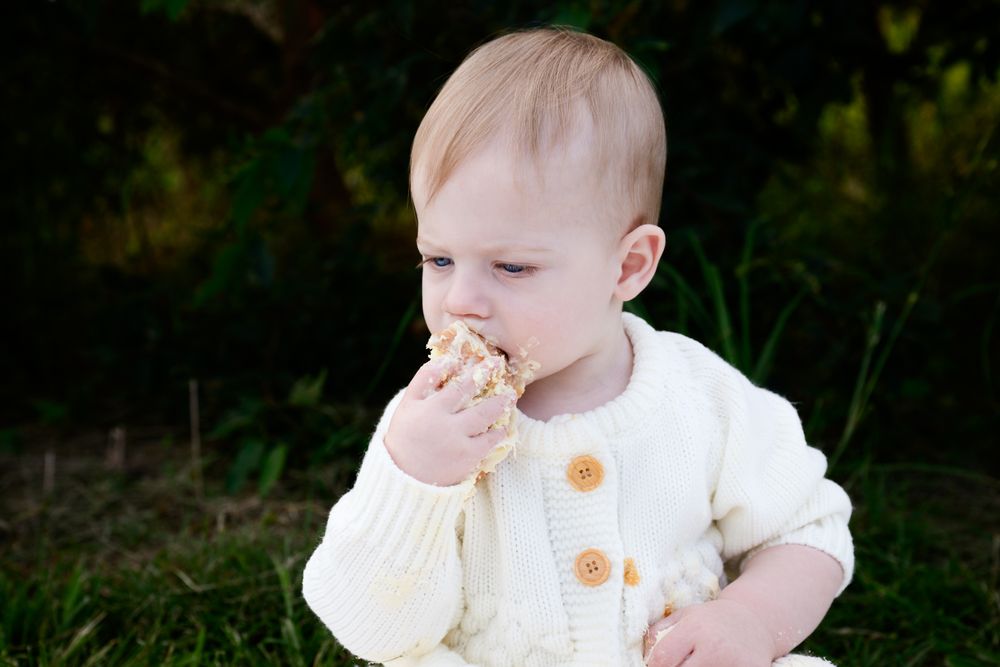 Small baby in cream colored clothing sits outdoors holding a cookie or biscuit snack in their hand.