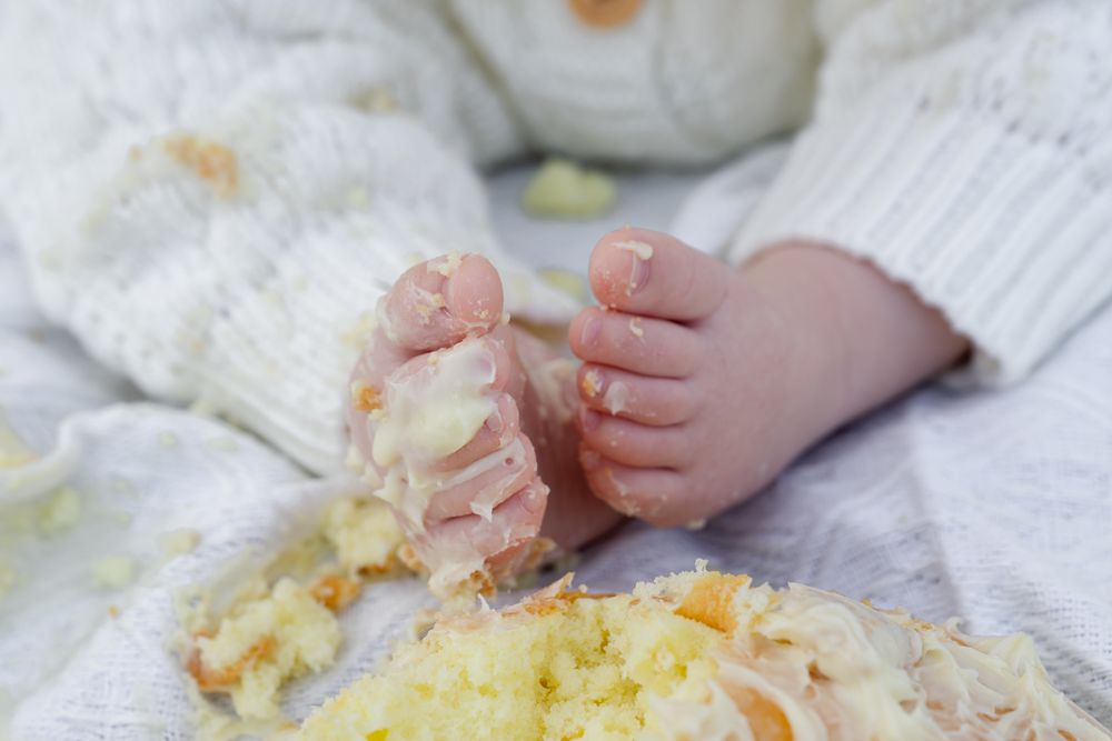 Baby feet next to smashed yellow birthday cake on white blanket during cake smash photo session.