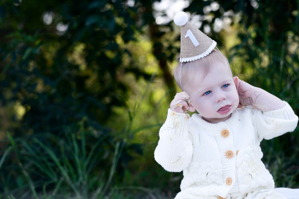 A small child wearing a white knitted sweater and party hat sits outdoors against a green natural background.