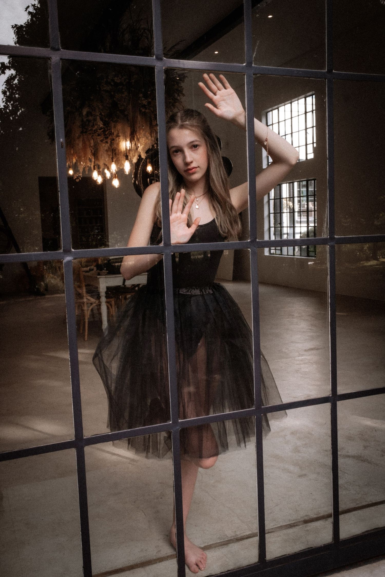 Teenage girl in black tulle dress standing behind metal window frame in moody industrial space.