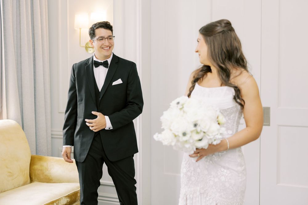 A couple share a moment during their first look, with the bride holding a white bouquet in an elegant indoor setting.
