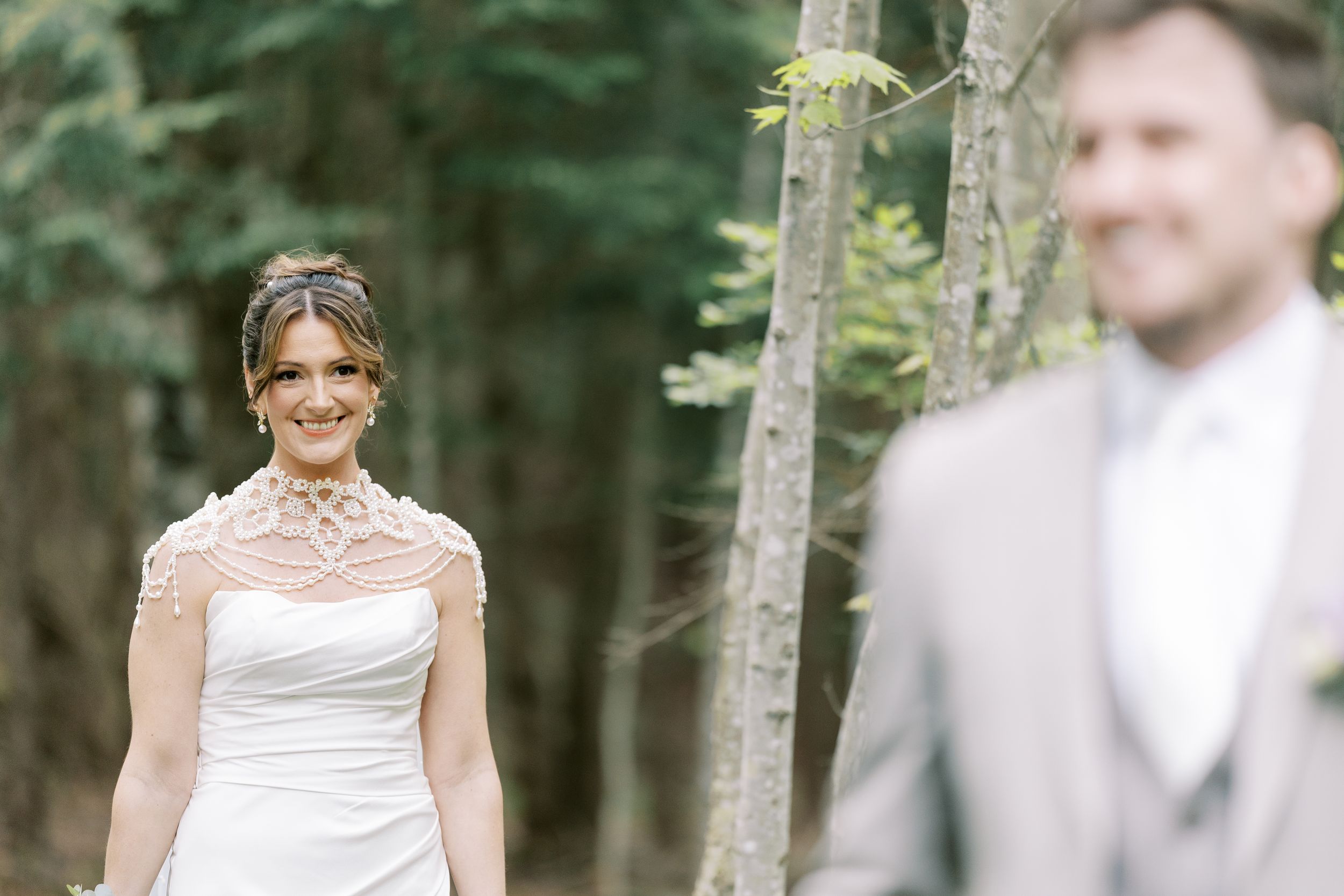 A romantic first look moment in the woods between a bride in white dress and groom in a grey suit.