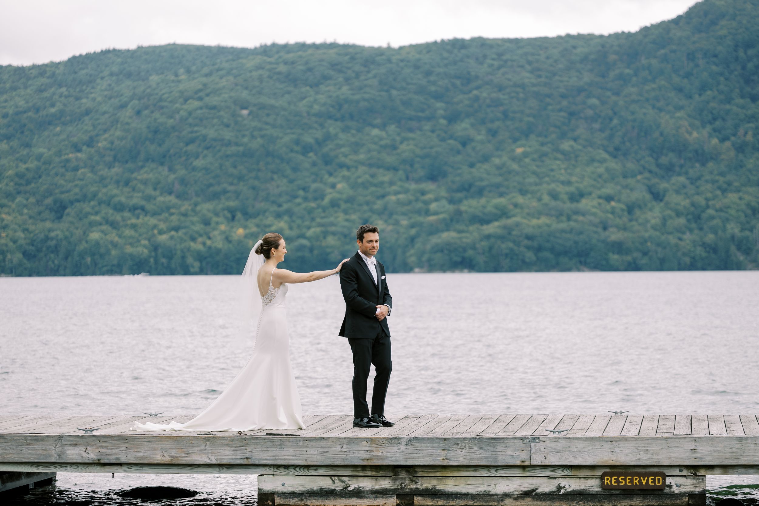 A couple stands on a scenic lakeside dock surrounded by mountains in wedding attire.