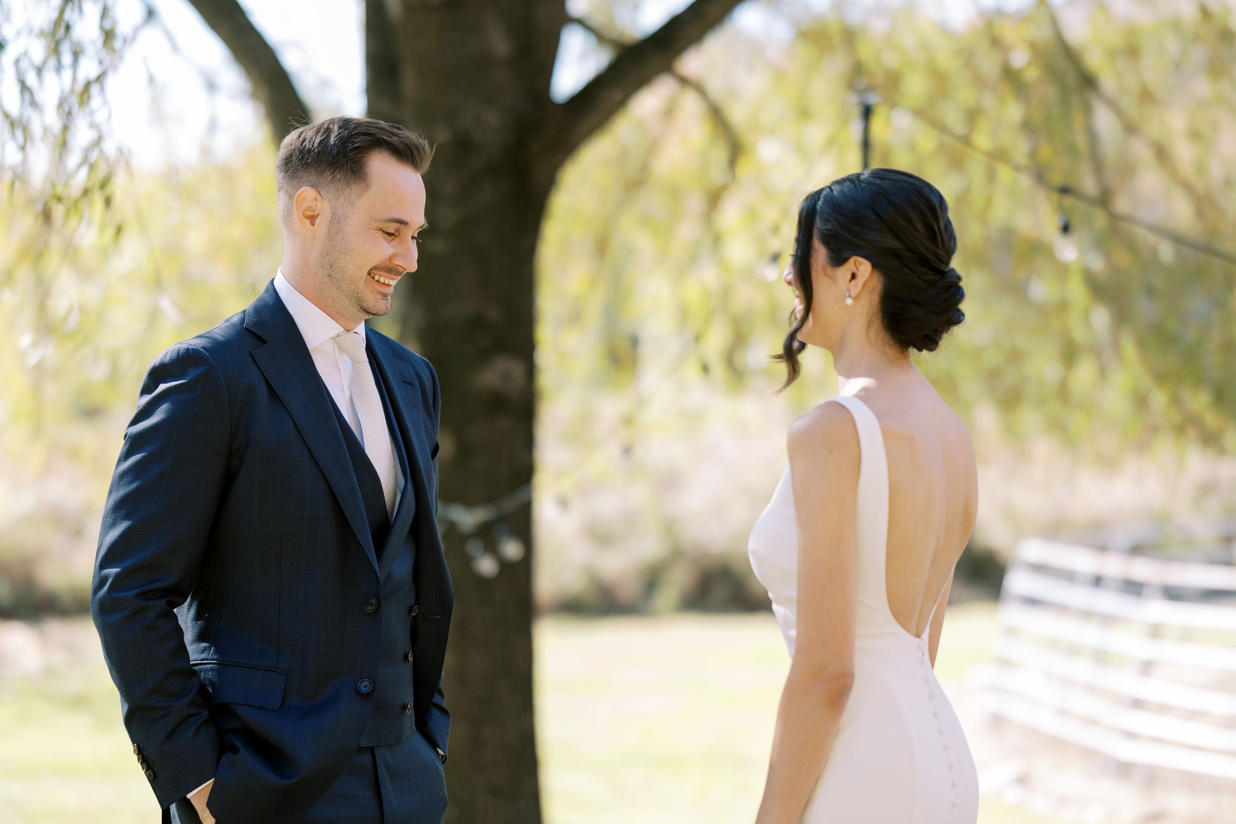 A couple wearing formal wedding attire smiles while walking together outdoors in a park setting.
