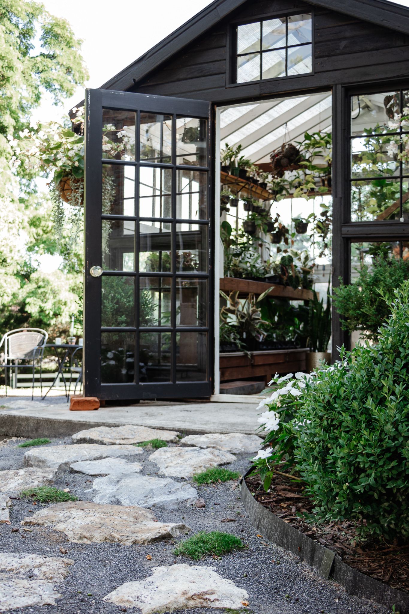 Black greenhouse with open doors, filled with lush greenery and hanging plants, surrounded by a stone path and garden.