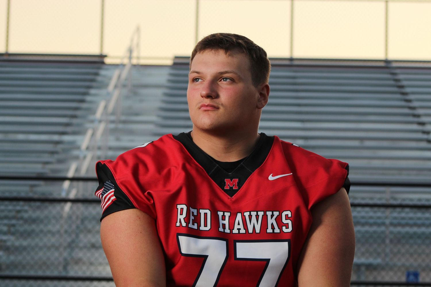 A football player wearing a red Red Hawks jersey number 77 stands in front of bleachers during golden hour.