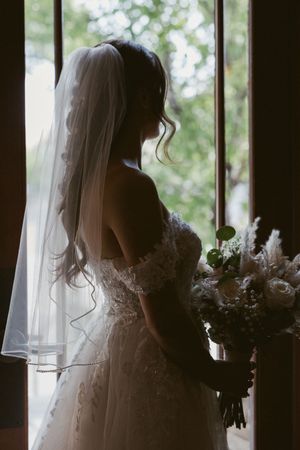 Silhouette of a bride in a lace wedding dress holding a bouquet while standing by a window.