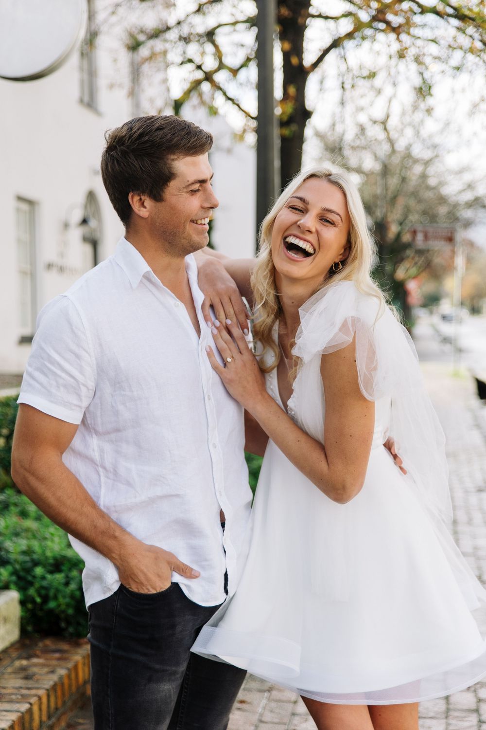 A couple laughs together during an intimate outdoor photoshoot, wearing white and black attire.