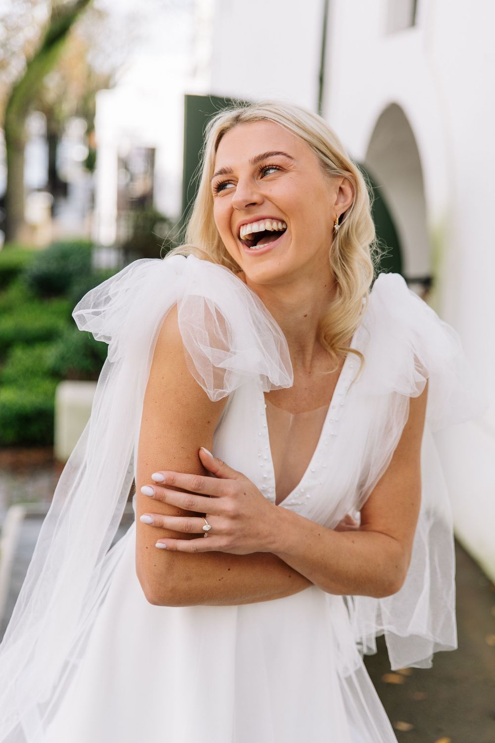 A bride in a white wedding dress with delicate tulle details laughs joyfully outdoors on her wedding day.