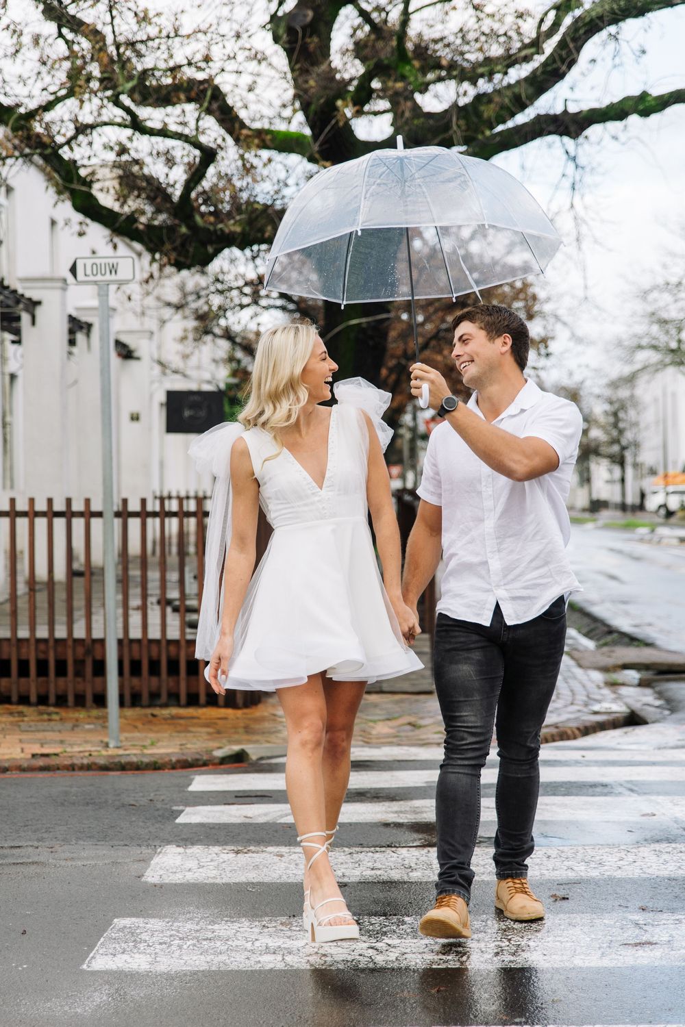 A couple shares a romantic moment under clear umbrellas on a rainy street with tree branches above.