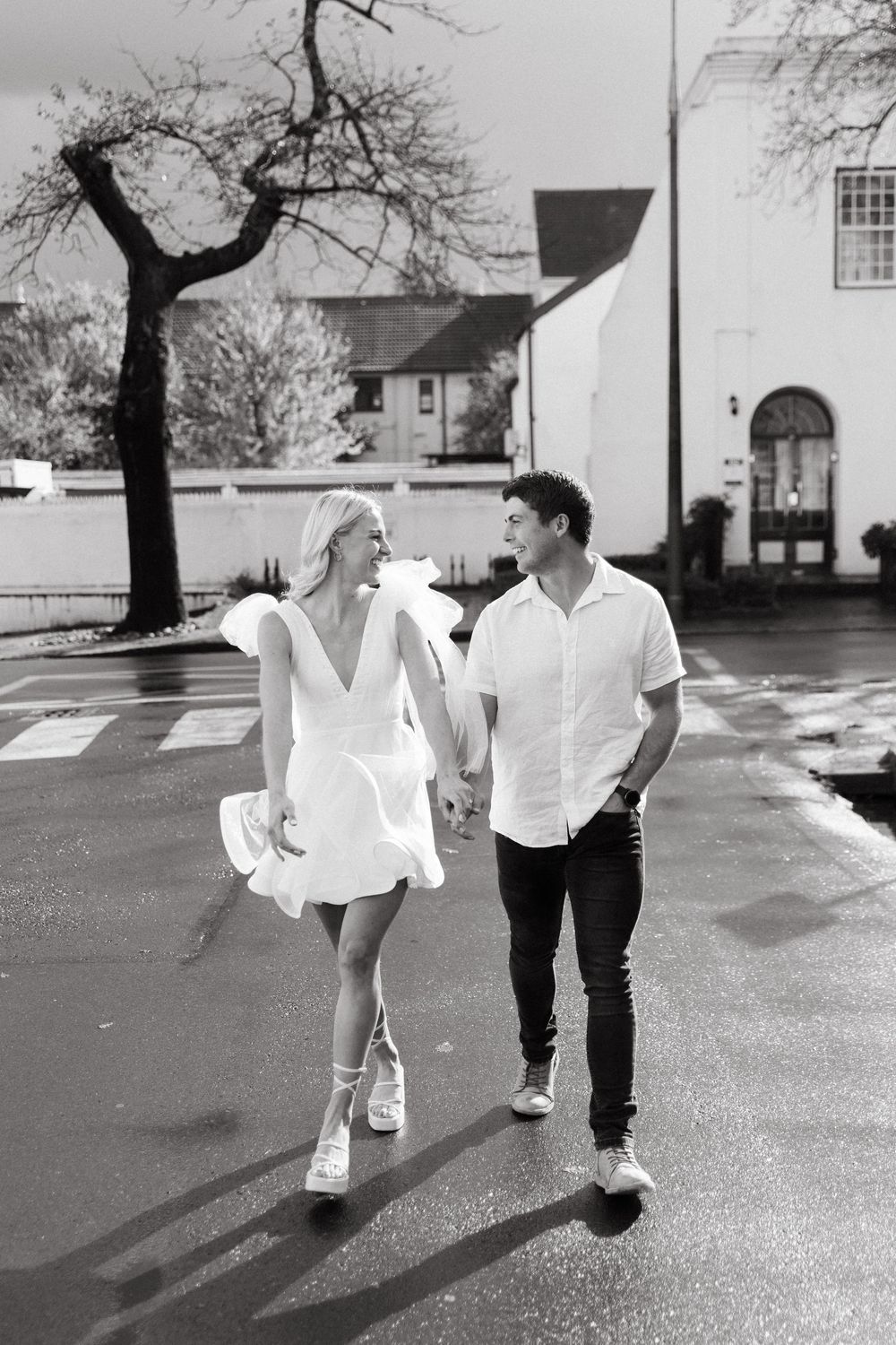 A romantic couple wearing white outfits dancing playfully on a street near trees and buildings in black and white.