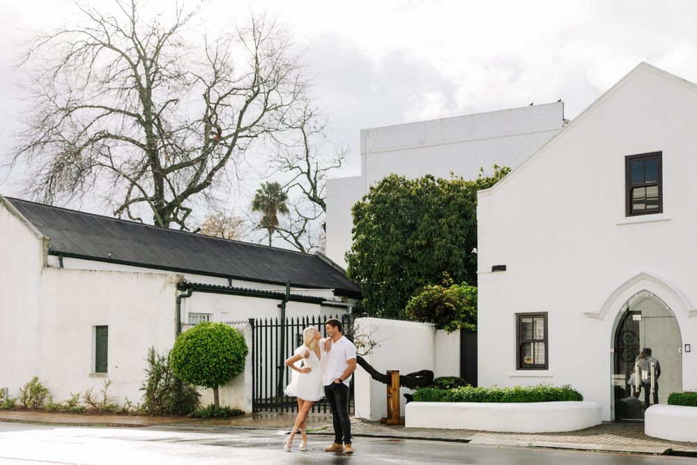A couple shares a romantic moment under an umbrella outside a white Mediterranean-style building with arched doorways.