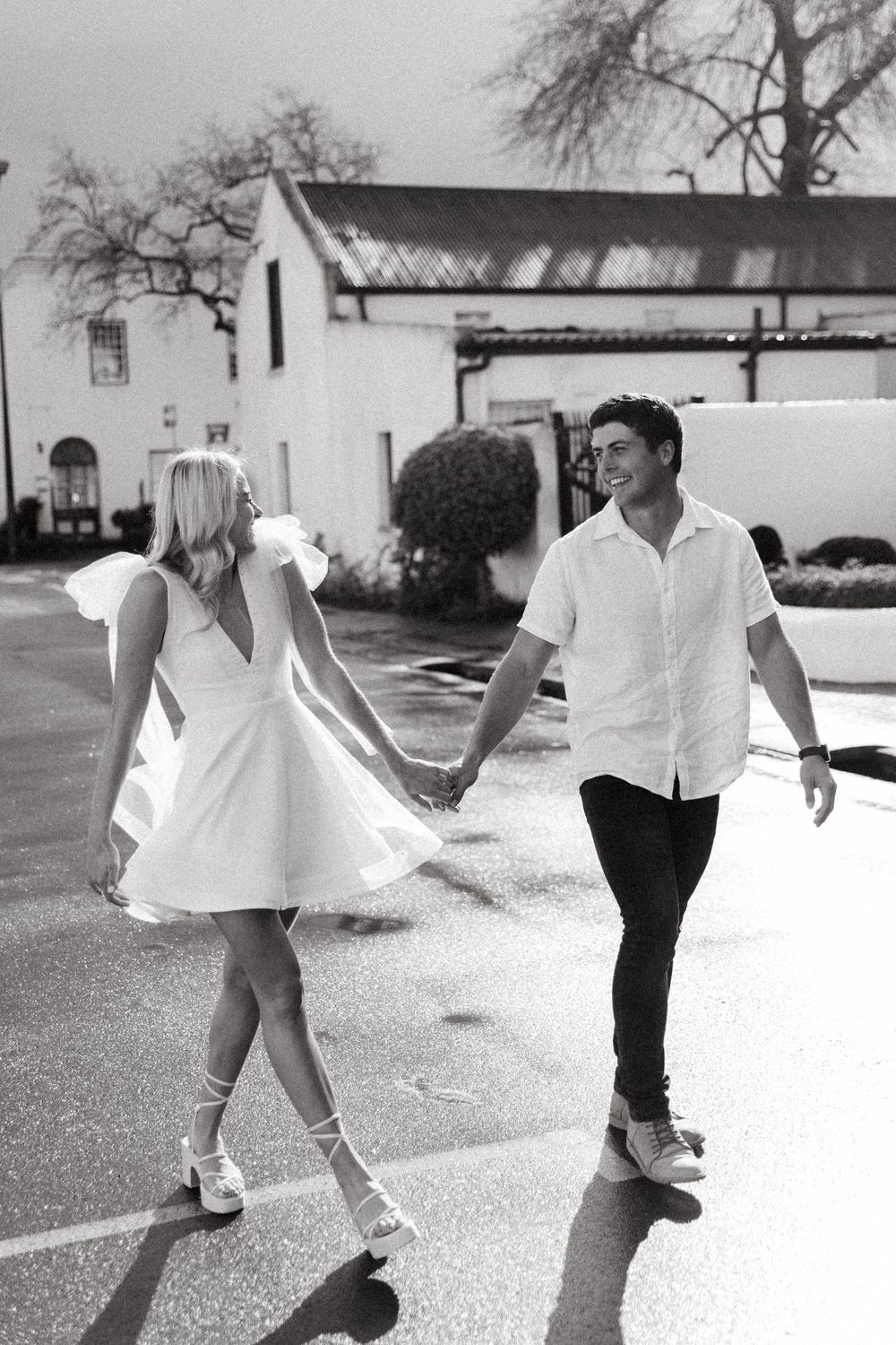 A couple in black and white dancing playfully outdoors, with her white dress twirling in front of a mission-style building.