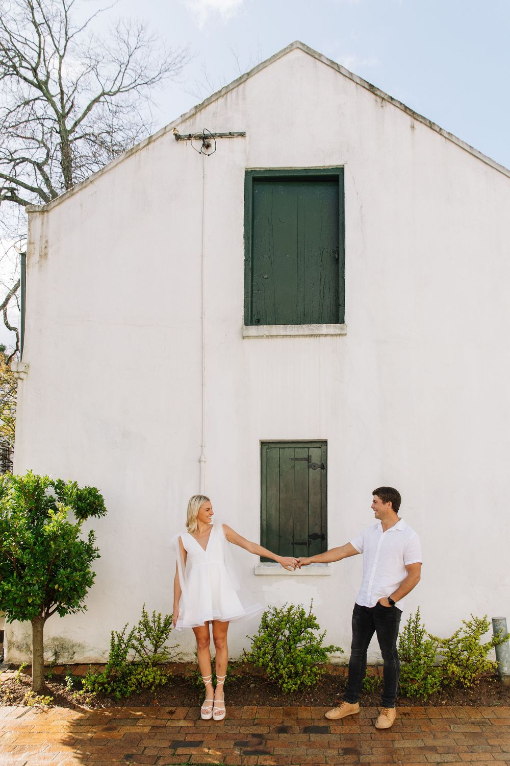 A couple in white outfits posing playfully against a white building with green shutters and doors in outdoor setting.