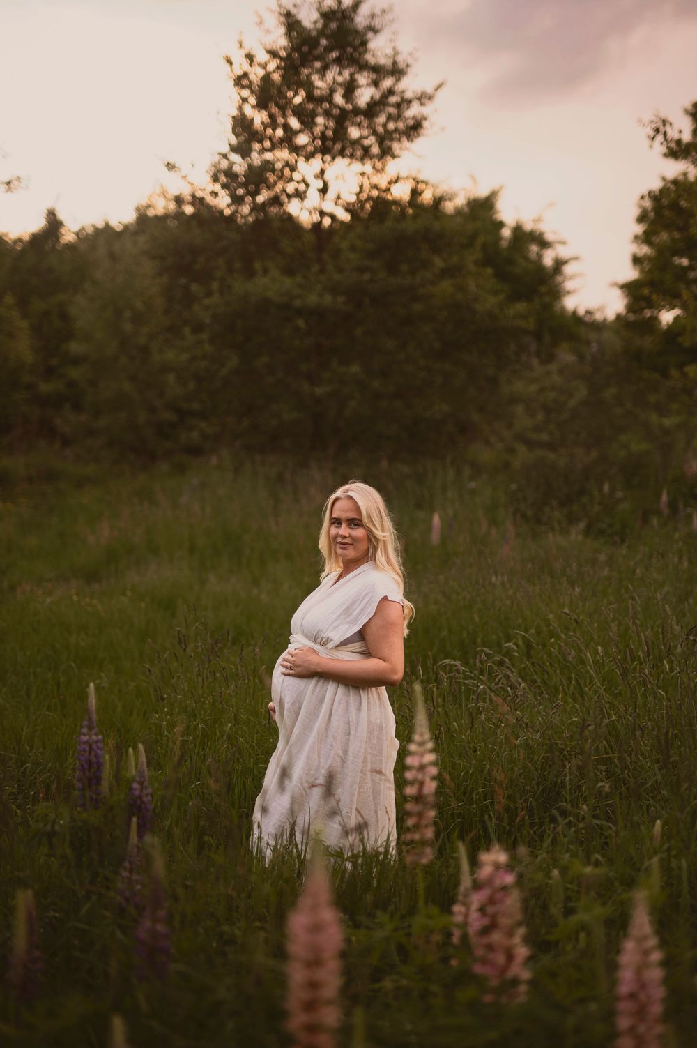 A pregnant woman in a white dress stands in a field of lupine flowers at sunset.