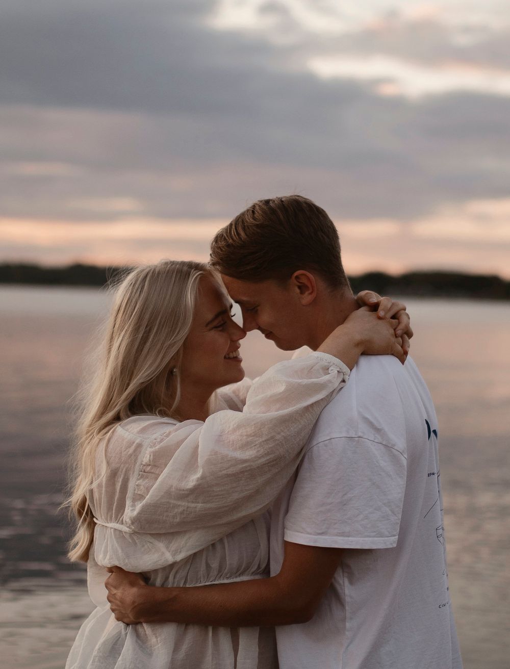 A romantic couple embraces by a serene lake during sunset, with dreamy clouds reflected in the tranquil water.