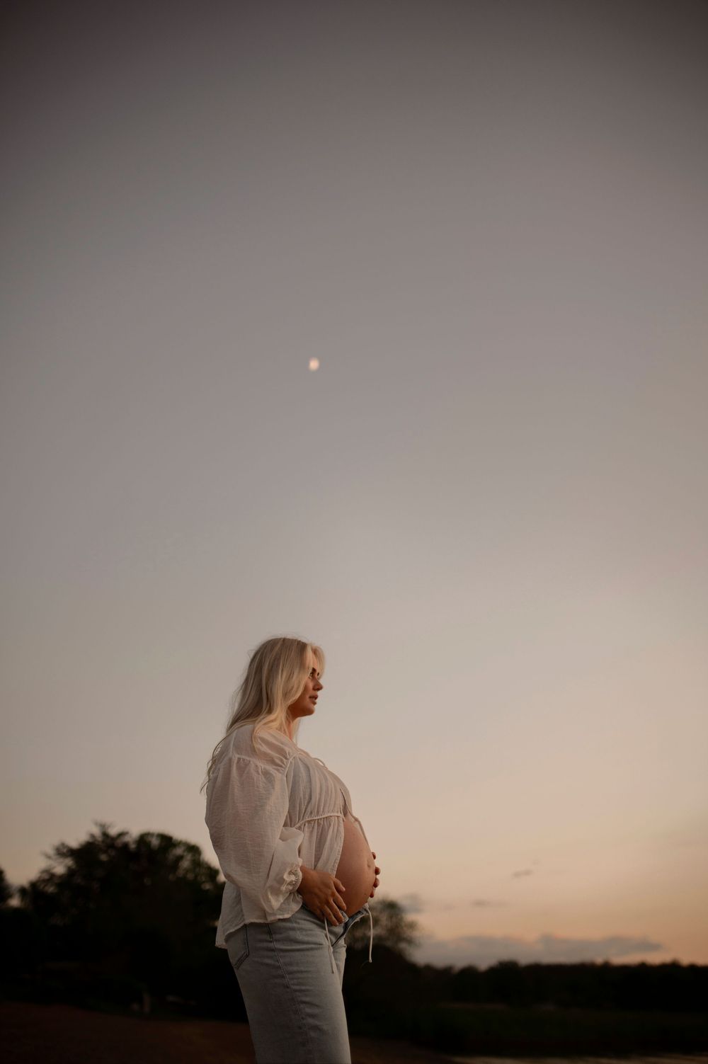 A pregnant figure stands in profile against a dusky sunset sky with a small moon visible overhead.