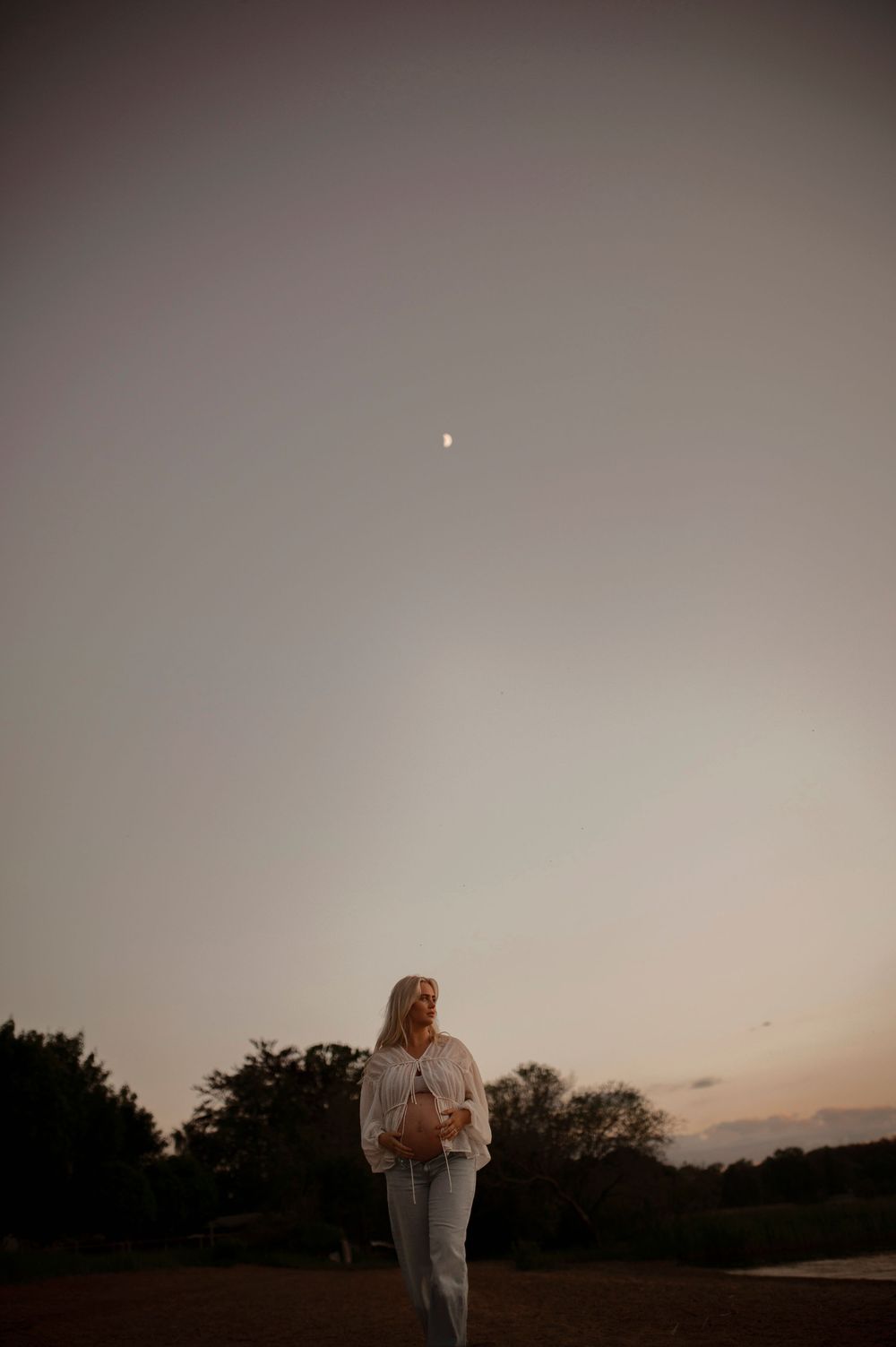 A silhouette of a person stands in a serene field at dusk with a crescent moon visible in the hazy evening sky.