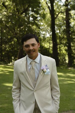 Groomsman in a cream colored suit with boutonniere poses in a sunlit wooded setting.