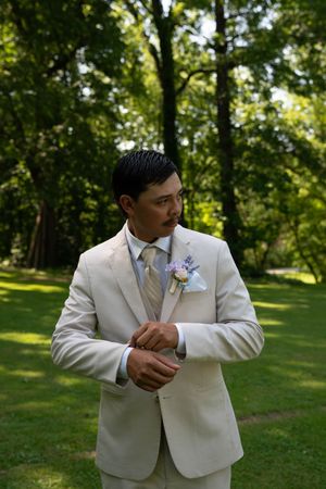 Groomsman adjusts his tie while standing in a forest clearing wearing a light colored suit.