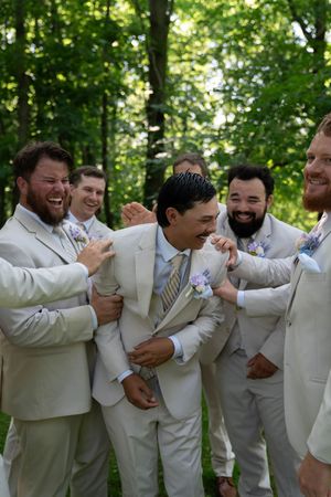 Wedding party in cream suits laughing and embracing in a playful group photo outdoors.