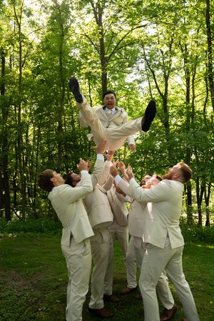Groomsmen in matching cream suits toss someone in the air during a fun wedding photo.