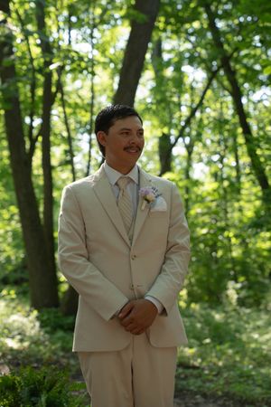 Two groomsmen in cream colored suits stand together in a sunlit forest setting.
