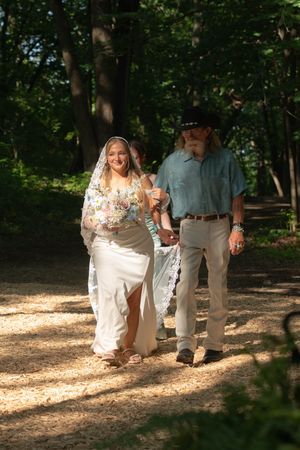 A couple walks together on a sunlit forest path during an outdoor wedding ceremony.