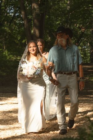 Wedding party members walk along a wooded trail dappled with sunlight.