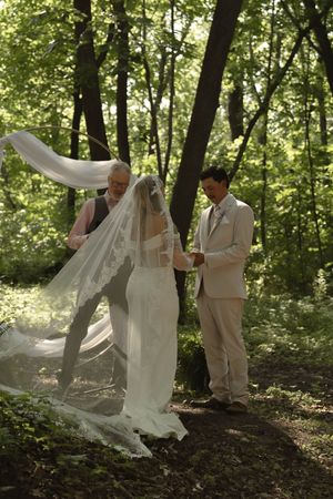 Wedding party poses with flowing veil in a sunlit forest setting.