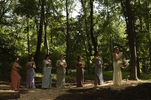 A wedding ceremony takes place in a sunlit wooded area with guests gathered around.